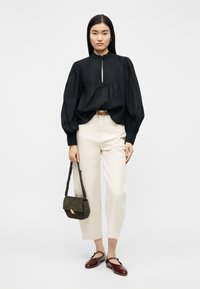 Γυναίκα που φοράει μαύρη μπλούζα με μακριά μανίκια και φουσκωτά μανίκια, εκρού κατεργασμένο παντελόνι, καφέ μπαλαρίνες και κρατάει μια μικρή σκούρα πράσινη τσάντα χειρός.