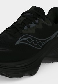 Chaussure de course athlétique noire avec tige en mesh, lacets noirs et logo gris sur le côté, inclinée pour montrer les détails avant et latéraux.