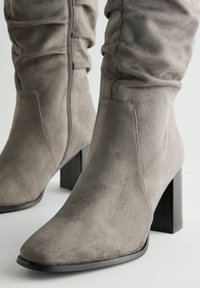 Graue Wildleder-Stiefeletten mit spitzem Zehenbereich, strukturierter Form und klobigem schwarzen Absatz; Merkmale sind ein seitlicher Reißverschluss und eine weiche Textur.
