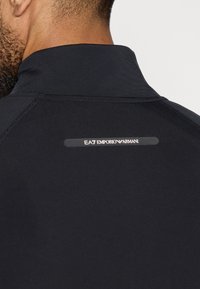 Jachetă sport neagră cu guler înalt, textură netedă și un logo reflectorizant EA7 Emporio Armani pe spate.