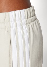 Beige joggebukser med ribbestrikket liv, med tre vertikale hvite striper langs sidene og et mykt, teksturert materiale.
