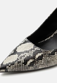 Chaussure à talon haut noire et blanche à bout pointu avec motif peau de serpent et finition brillante sur fond uni.