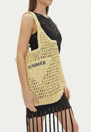 Mujer con vestido negro sosteniendo un bolso tote tejido de color beige con el texto "SUMMER" bordado en el bolsillo delantero.