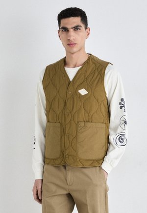 Quilted olive green vest with a V-neck design, two large front pockets, and a label detail on the left side. Worn over a white long-sleeve shirt.