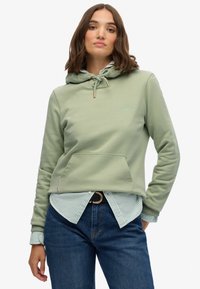 Sudadera verde claro con un bolsillo delantero, capucha ajustable con cordón y logotipo bordado. Combinada con una camisa a rayas azul y blanca y jeans.