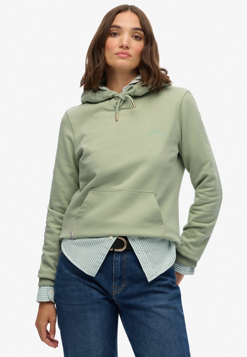 Sudadera verde claro con un bolsillo delantero, capucha ajustable con cordón y logotipo bordado. Combinada con una camisa a rayas azul y blanca y jeans.