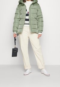 Padded green jacket with a hood, zip closure, and horizontal stitching. Paired with cream pants and white sneakers, holding a black handbag.