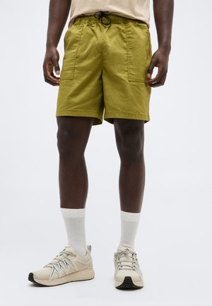 Άνδρας που φοράει χακί σορτς με μπροστινές τσέπες, λευκές κάλτσες μέσης γάμπας και μπεζ παπούτσια τρεξίματος, σε λευκό φόντο.