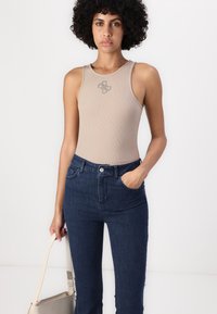 Top sin mangas beige con canalé, cuello redondo y un sutil detalle de logo, combinado con unos jeans de tiro alto en azul oscuro. La modelo sostiene un bolso claro.
