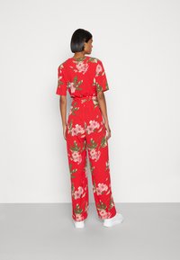 Macacão floral vermelho com mangas curtas, cintura cingida e pernas largas. Apresenta flores cor-de-rosa e folhas verdes, combinado com sapatos brancos.