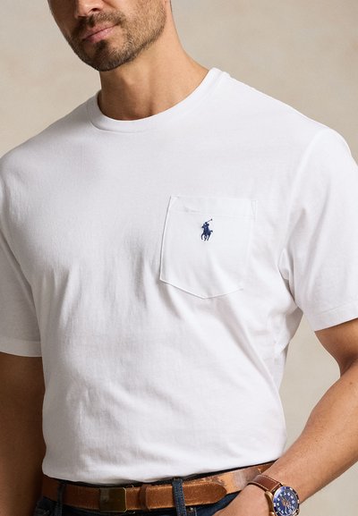 Mand iført hvid kortærmet t-shirt med rund hals, brystlomme og blåt broderet hestelogo, brun bælte og armbåndsur.