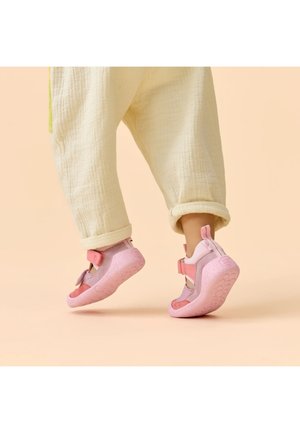 Peuter draagt gebroken witte broek en roze zachte schoenen, staat op haar tenen tegen een beige achtergrond.