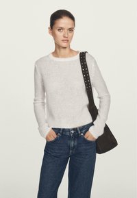 Donna con le mani in tasca, che indossa un maglione lavorato a maglia bianco, jeans blu scuro e una borsa a spalla nera con borchie, su uno sfondo neutro.