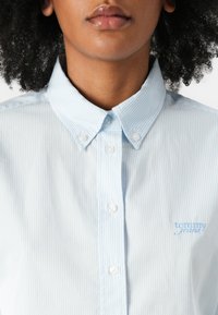 Light blue striped button-up shirt with a collar, featuring white buttons and a small embroidered logo in light blue on the left side.