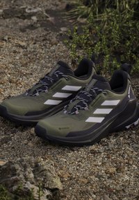 Zapatos de senderismo verde y negro con rayas blancas, parte superior texturizada, suela resistente y cordones con patrón, con la marca GORE-TEX en el lateral.