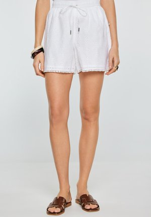 Białe szorty z tkaniny z ażurowym wzorem, z paskiem ściągającym w talii i lamówką z pomponami, noszone z brązowymi sandałami na płaskiej podeszwie na stojącej osobie.