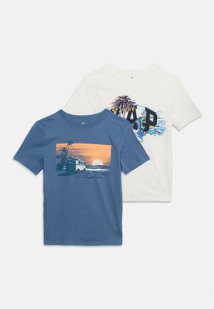 Kék póló naplementés tengerparti és kisteherautó mintával, fehér póló pálmafákkal, óceánnal és nagy GAP felirattal.