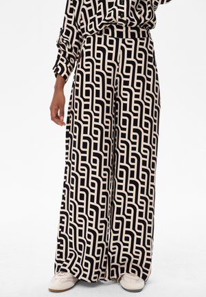 Spodnie o szerokich nogawkach z czarno-beżowym geometrycznym wzorem przypominającym labirynt, noszone z dopasowanym topem o tym samym wzorze i białymi trampkami.