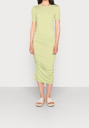 Femme portant une robe vert clair jusqu'au genou avec des rayures blanches ondulées et des sandales beige ouvertes aux orteils, debout devant un fond uni.