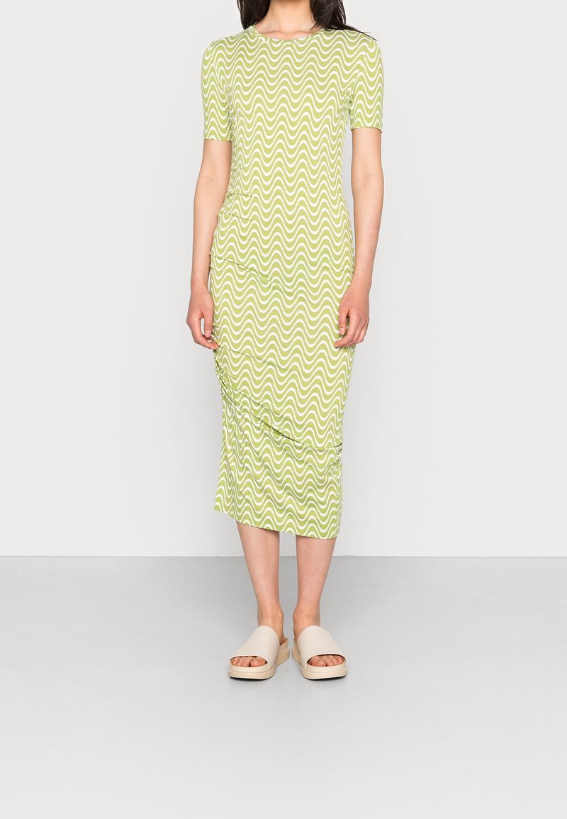 Femme portant une robe vert clair jusqu'au genou avec des rayures blanches ondulées et des sandales beige ouvertes aux orteils, debout devant un fond uni.