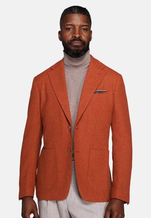 Man wearing a rust orange blazer over a beige turtleneck and light gray pants, standing against a plain white background.