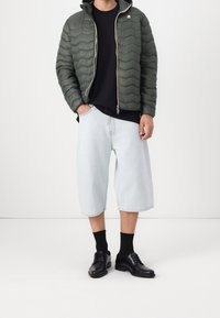 Homme portant une veste matelassée vert olive, une chemise noire, un short en denim bleu clair à mi-genoux, des chaussettes noires et des chaussures habillées en cuir noir.