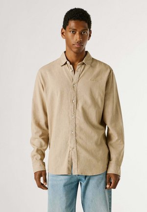 Giovane uomo con camicia beige a maniche lunghe abbottonata e jeans azzurri, in piedi davanti a uno sfondo bianco uniforme.