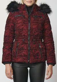 Kobieta w czerwonej, wzorzystej zimowej kurtce z czarnym futrzanym kapturem i zapinanymi kieszeniami, w połączeniu z czarnymi spodniami.