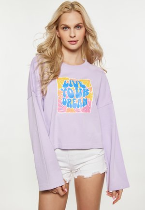 Lyshåret kvinde bærer en lyse lilla langærmet skjorte med teksten "Live Your Dream" og motiver, kombineret med hvide frynser-shorts.