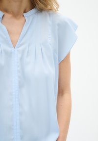 Blusa azzurro chiaro con maniche corte a sbuffo, dettagli plissettati sul colletto e una texture di tessuto liscia.