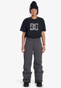 Grå snöbyxor med avslappnad passform, i kombination med en svart T-shirt med en silverfärgad DC-logotyp. Avslutat med en svart mössa.