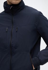 Μπλε ναυτικό μπουφάν με φερμουάρ, κατασκευασμένο από λείο ύφασμα, με δύο τσέπες στο στήθος με φερμουάρ, ελαστικά μανίκια και ψηλό γιακά.