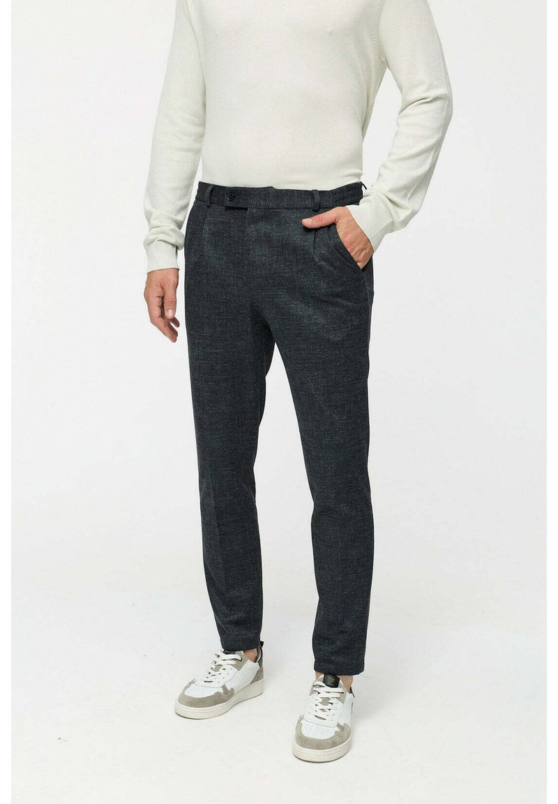 Dunkelgraue, strukturierte Hose mit einer taillierten Passform, ausgestattet mit seitlichen Taschen und flachem Bund. Getragen mit einem weißen Langarmoberteil und Turnschuhen.