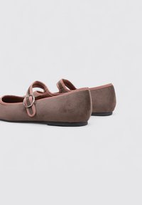Zapatos planos de ante marrón con puntera redondeada, correa en el tobillo y detalle de hebilla metálica. Presentan una textura suave y suela plana negra.