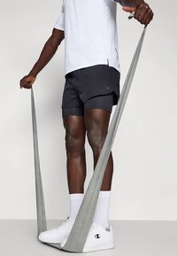 Egy fehér sportcipőt, fekete rövidnadrágot és fehér zoknit viselő személy egy ellenállás szalagon áll, és mindkét kezével felfelé húzza.