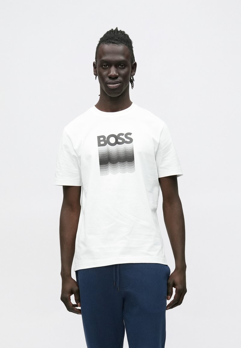 Mlajši moški s spetimi lasmi, oblečen v belo majico s tekstom "BOSS" v črno-sivem gradientu, kombinirano z mornarsko modrimi hlačami, stoji pokončno.