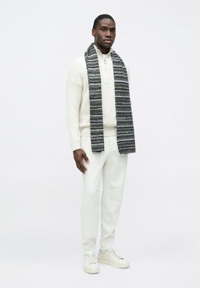 Πλεκτό εκρού πουλόβερ με φερμουάρ στον γιακά, συνδυασμένο με λευκό παντελόνι και ανοιχτό γκρι παπούτσια. Μια ριγέ γκρι εσάρπα είναι περασμένη γύρω από το λαιμό.
