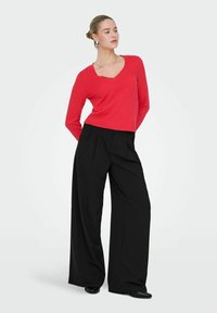 Femme portant un haut rouge à manches longues avec un col en V et un pantalon noir taille haute à larges jambes, debout les mains derrière le dos.