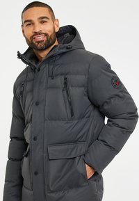 Mörkgrå puffjacka med huva, med dragkedje- och ficklockfickor, texturerad tyg och en logotypapplikation på vänstra ärmen.