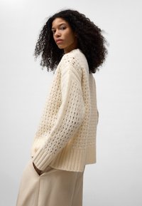 Creme gebreide trui met een getextureerd opengeweven ontwerp, geribde manchetten en zoom. Model draagt beige trousers. Monochroom achtergrond.