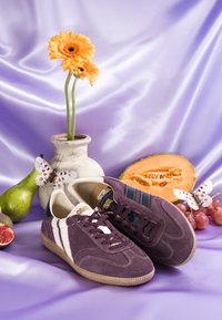 Baskets en daim violet avec des rayures blanches à côté d'un vase avec des fleurs orange, une moitié de melon, des raisins, une poire, une figue et des papillons décoratifs sur un tissu violet.