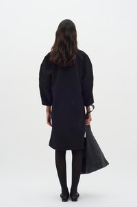 Schwarzes strukturiertes Kleid mit lockeren Ärmeln, in mittlerer Länge, getragen mit schwarzen Strumpfhosen und flachen Schuhen. Das Modell trägt eine große graue Tragetasche.