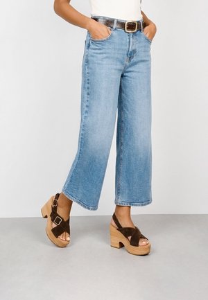 Mujer con vaqueros cortos de pierna ancha de color azul claro, cinturón marrón y sandalias marrones de plataforma con tacones de madera, de pie con las manos en los bolsillos.