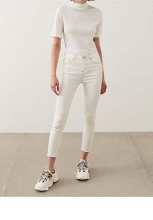 Persoon draagt strakke witte hoge taille jeans, wit kortärmlig topje en chunky witte sneakers met zwarte veters, staand op een grijze vloer.