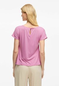 Top rosa a maniche corte con dettaglio a chiave sul retro, tessuto morbido e vestibilità rilassata, abbinato a pantaloni chiari.