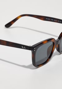 Tortoisepruunist Ray-Ban päikeseprillid tumevalgete neljkandiliste objektiividega, nähtava logoga raamimisel ja objektiivil, esitatud valgel taustal.