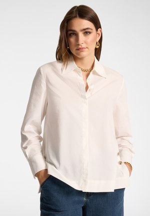 Femme aux cheveux bruns portant un chemisier blanc boutonné et un jean bleu, accessoirisée d'un collier chaîne en or, de boucles d'oreilles créoles et d'un bracelet.