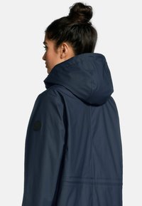 Marineblauwe waterdichte jas met een capuchon, gladde textuur en een cirkelvormig zwart logo op de linker mouw. Heeft een naaddetail op de taille.