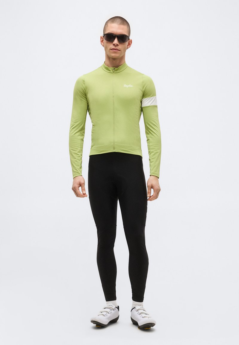 Mann trägt hellgrünes langärmliges Radtrikot, schwarze Radlerhose, weiße Radschuhe und schwarze Sonnenbrille, steht vor einfarbigem Hintergrund.