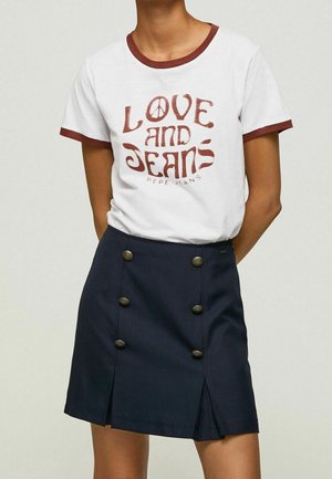 Persona che indossa una maglietta bianca con bordi marroni e testo "Love and Jeans", abbinata a una gonna blu navy con due file verticali di bottoni.
